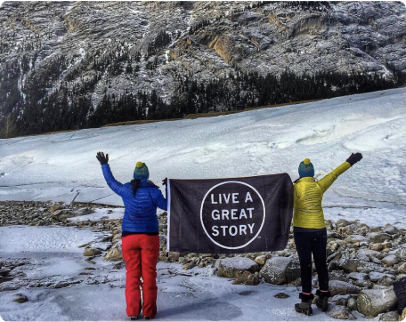 Two people in the snow holding up a LIVE A GREAT STORY Adventure Flag