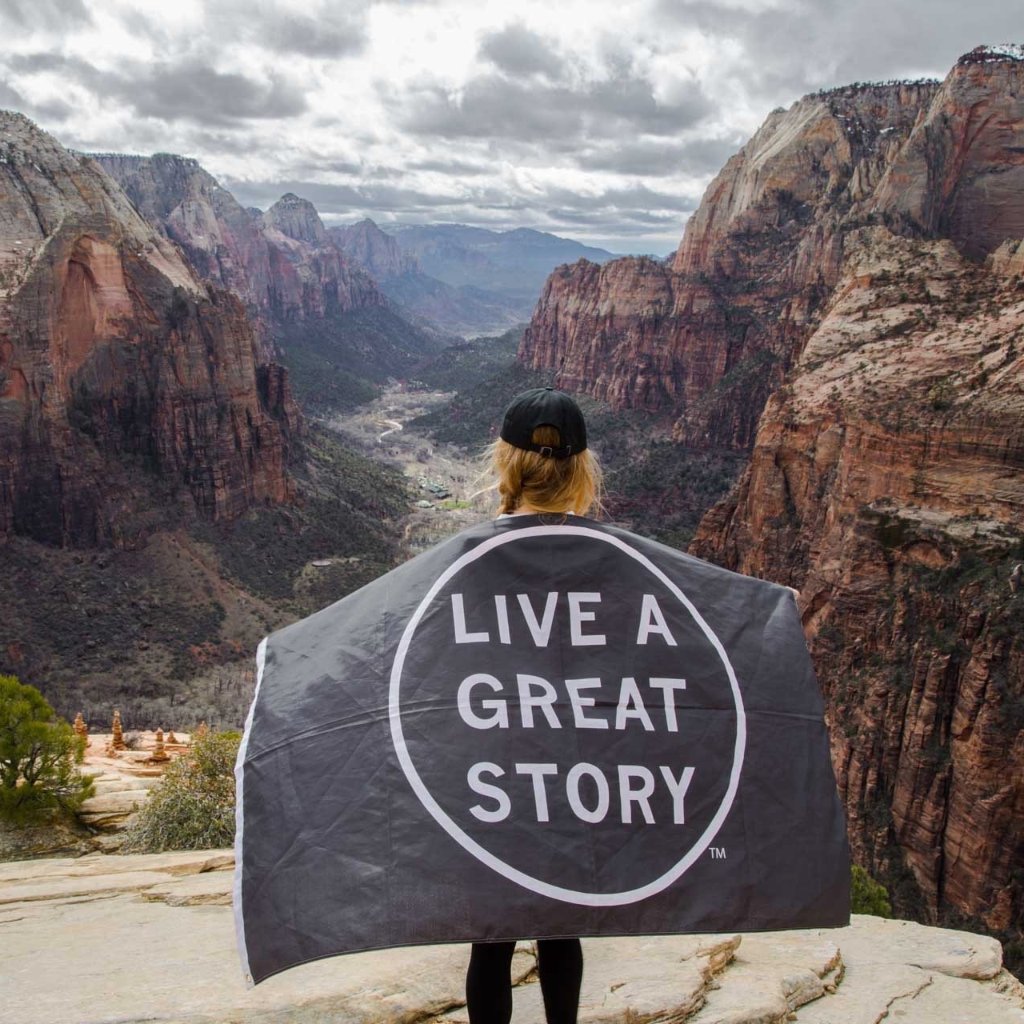 A person holding a black LIVE A GREAT STORY Adventure Flag at a scenic mountain valley