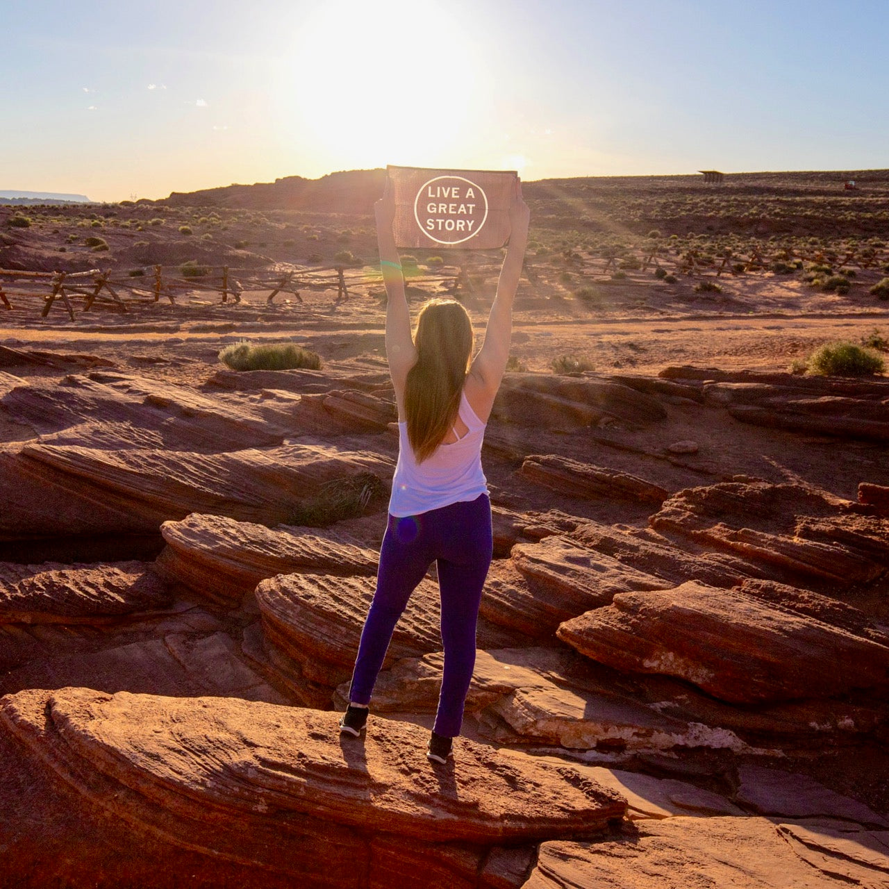 A woman holding up a black LIVE A GREAT STORY Mini Adventure Flag with a scenic outdoor view