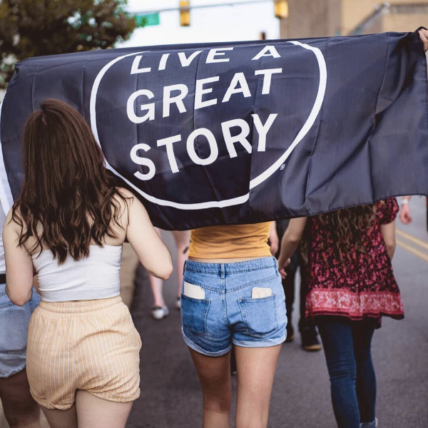 A group of people walking and holding a black LIVE A GREAT STORY Adventure Flag