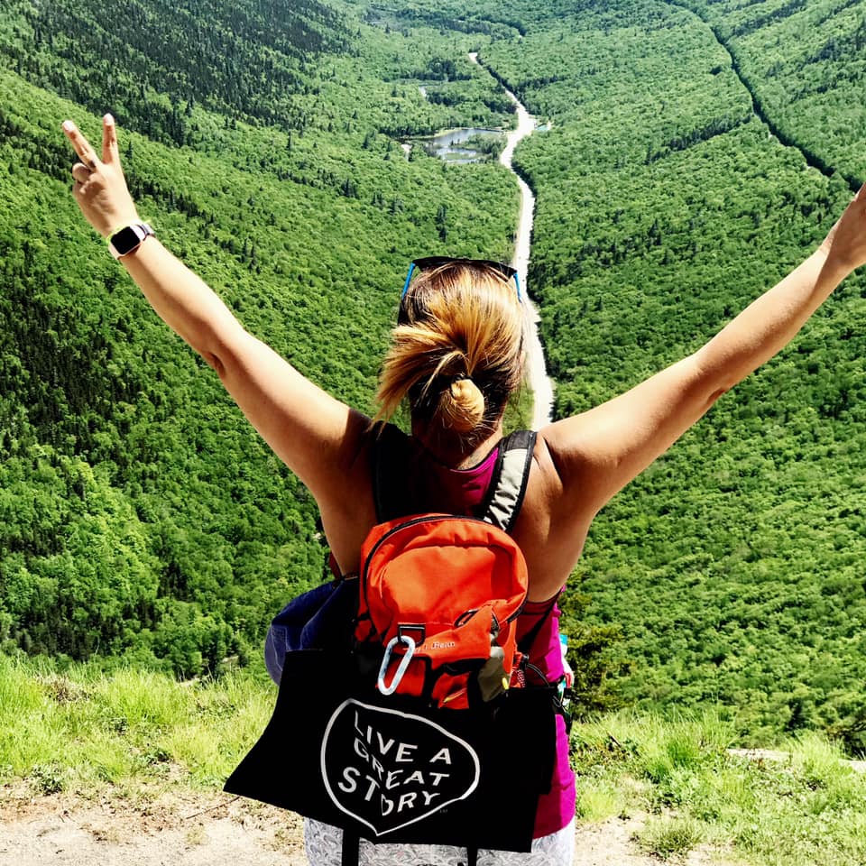 A with a black LIVE A GREAT STORY Mini Adventure Flag on their backpack at a scenic location