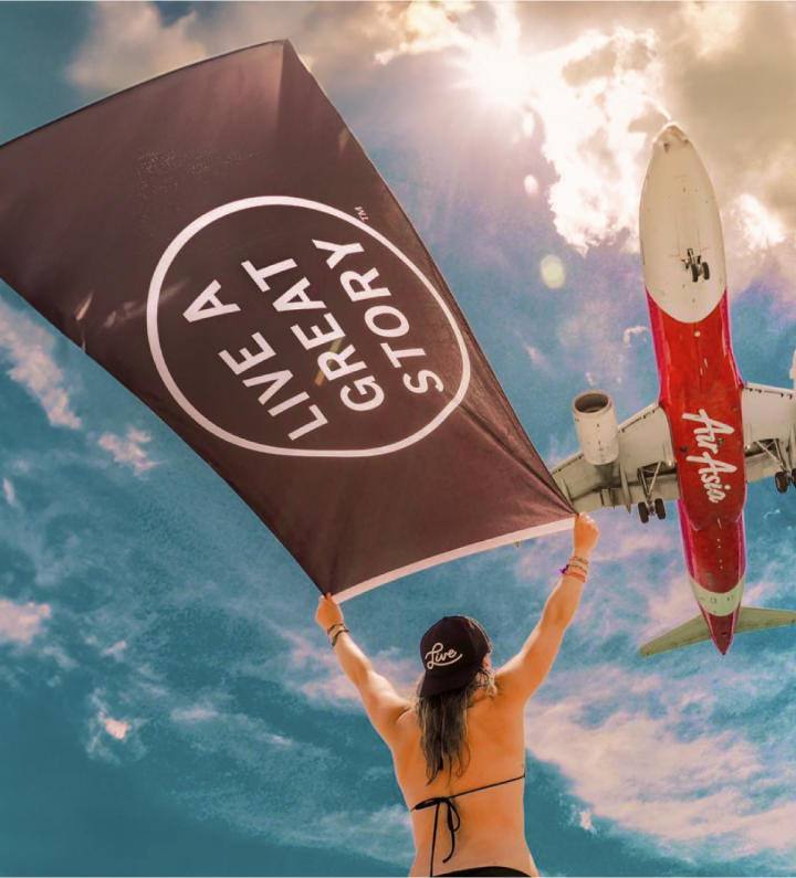 A woman holding up a LIVE A GREAT STORY Adventure Flag with an airplane flying above