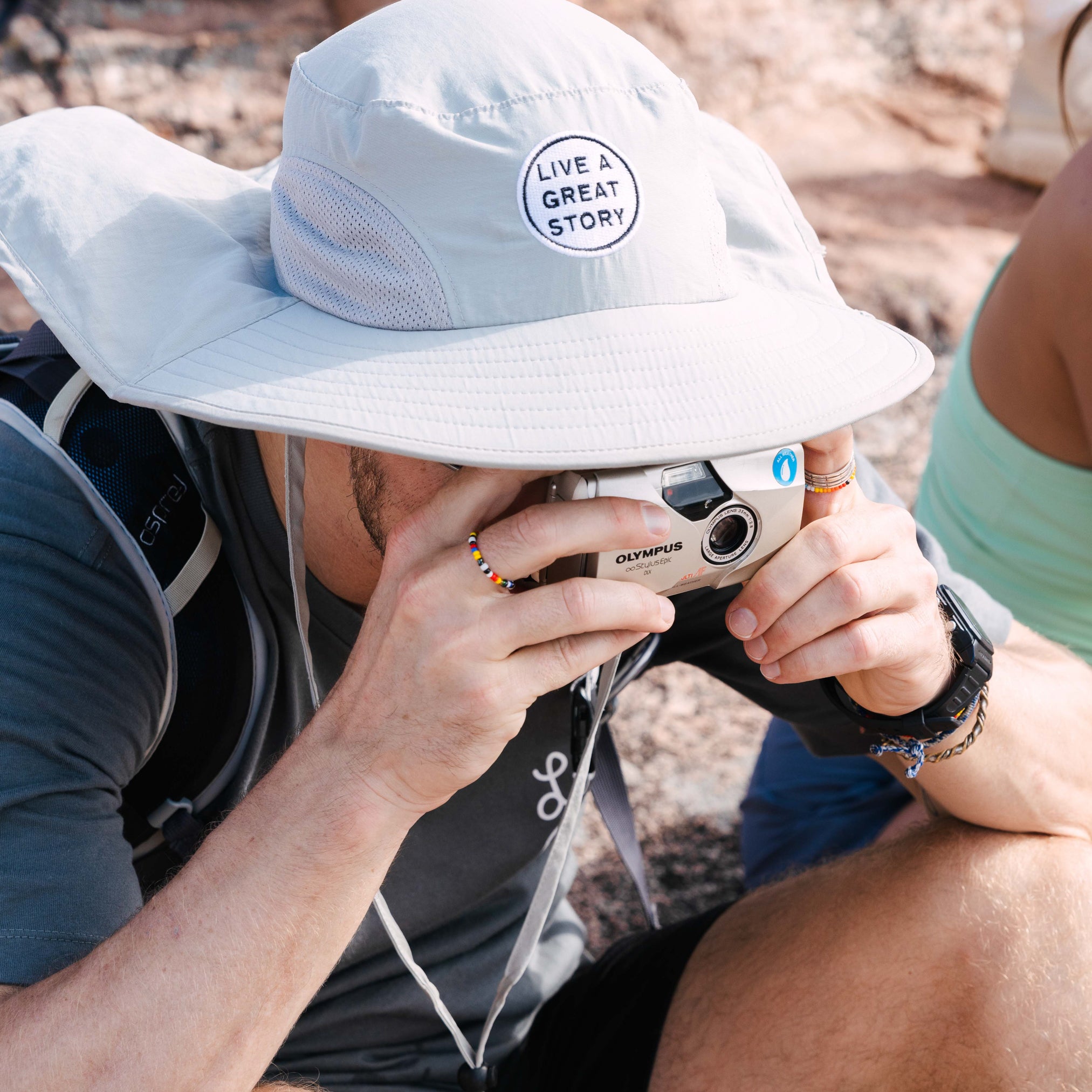 A man wearing a LIVE A GREAT STORY Adventure Hat while taking a photo