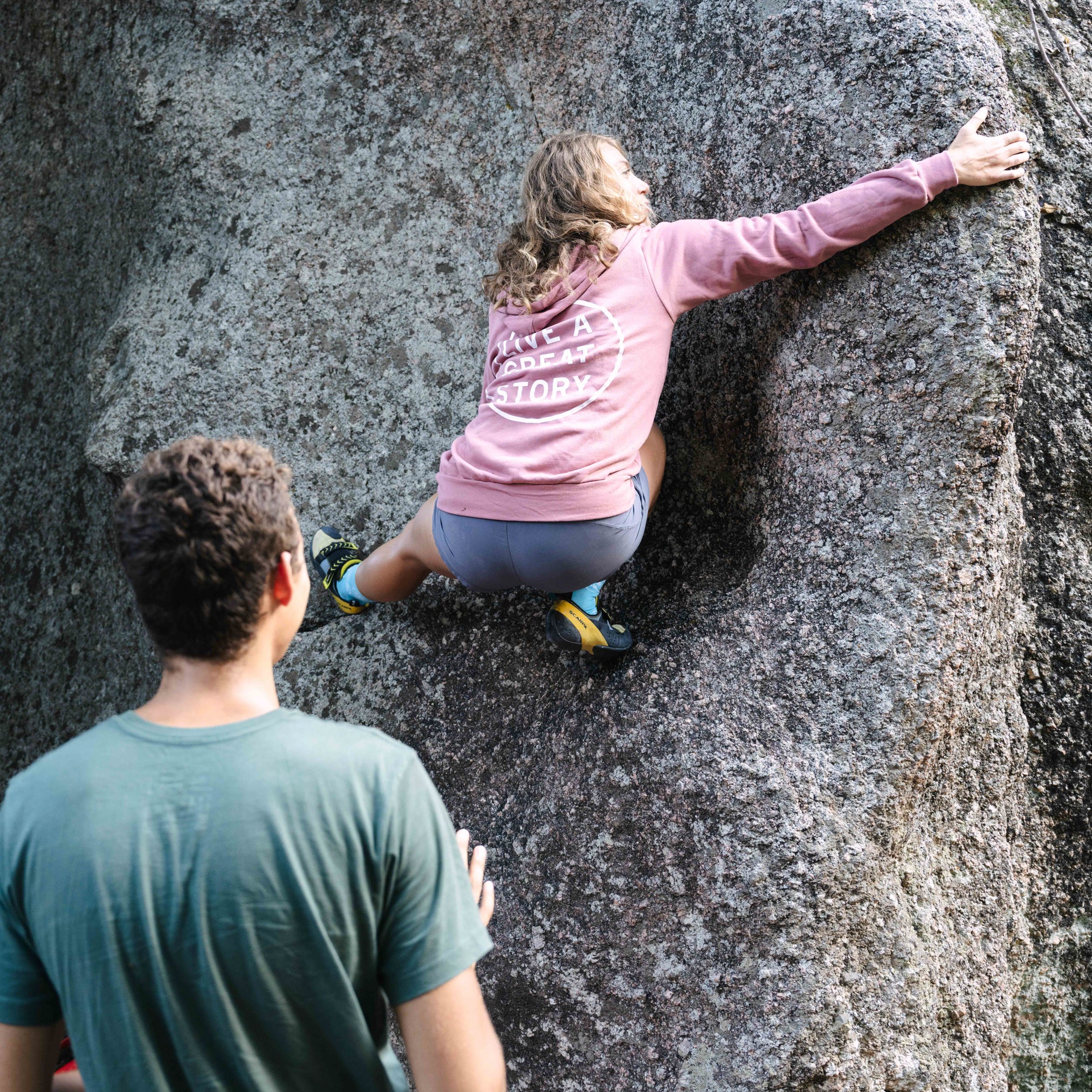 A person wearing a mauve LIVE A GREAT STORY zip up hoodie while climbing a rock