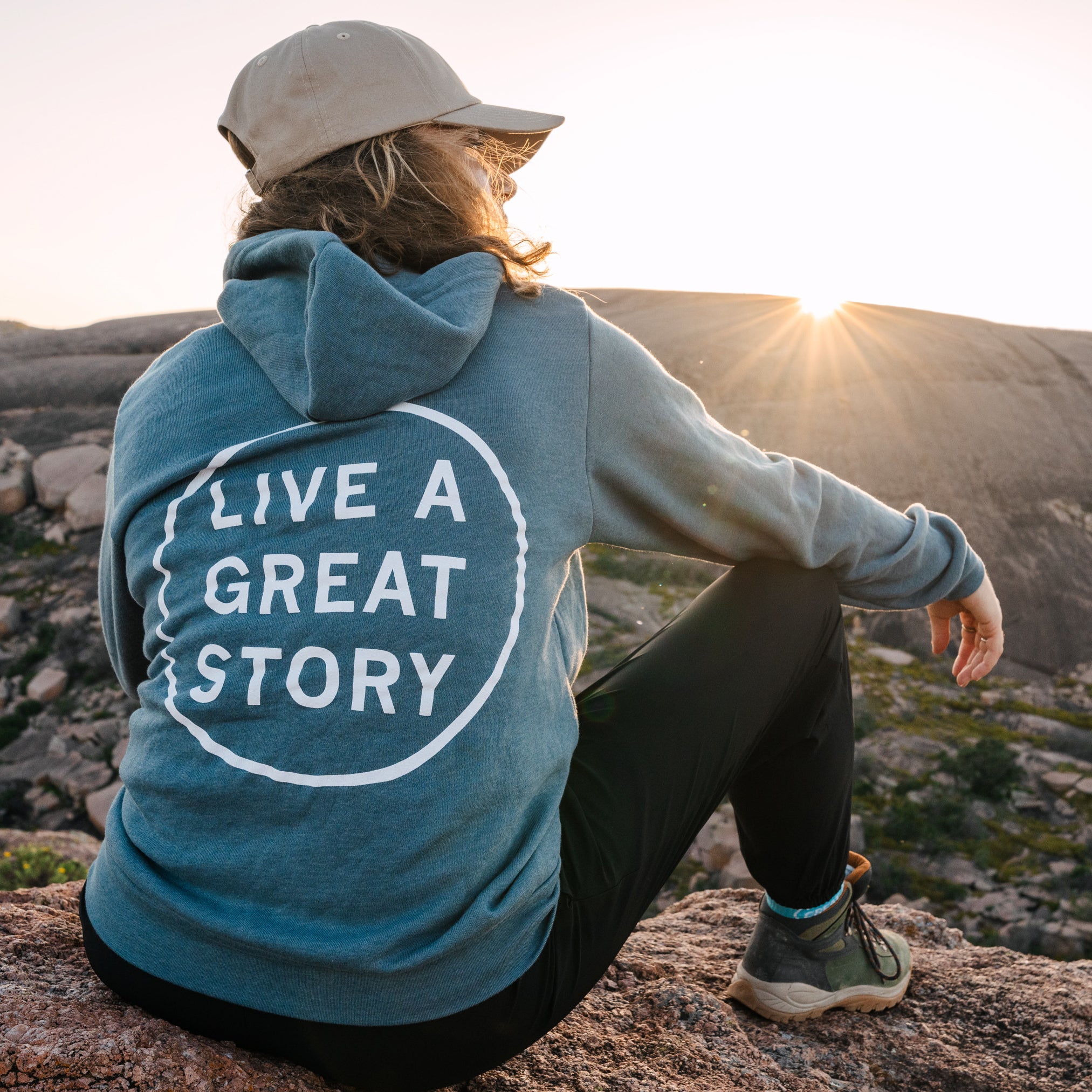 A person wearing a blue LIVE A GREAT STORY zip up hoodie while watching a sunset