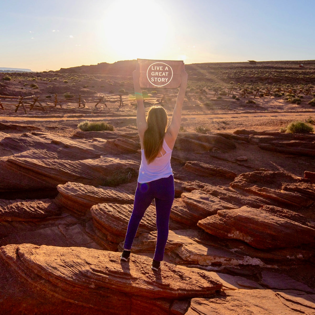 A woman holding up a black LIVE A GREAT STORY Mini Adventure Flag with a scenic outdoor view