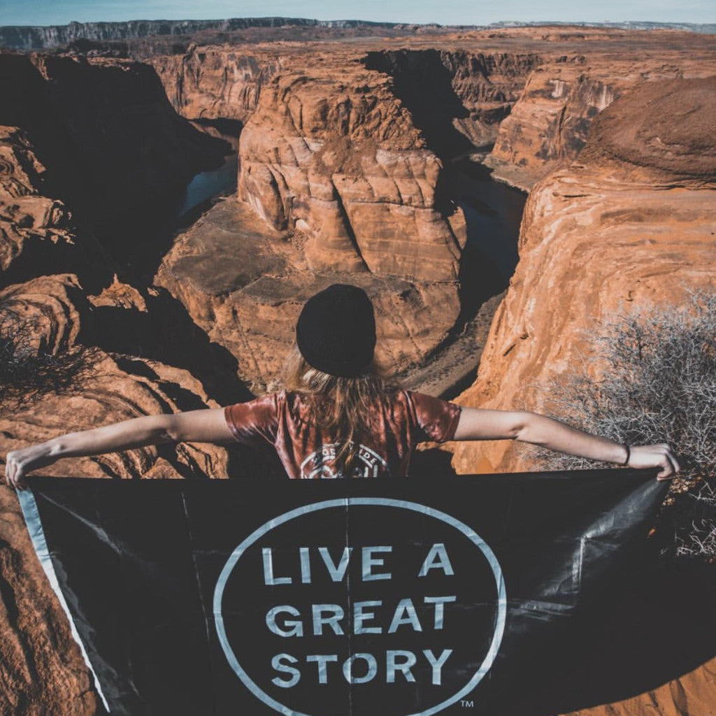 A person holding a black LIVE A GREAT STORY Adventure Flag at a scenic valley